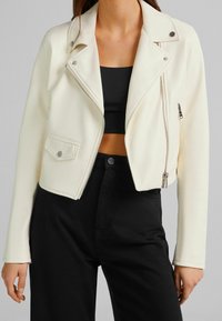 Kremowa krótka kurtka z ekologicznej skóry z srebrnymi zamkami błyskawicznymi, zatrzaskami i klasycznym motocyklistycznym krojem, zestawiona z czarnym topem.