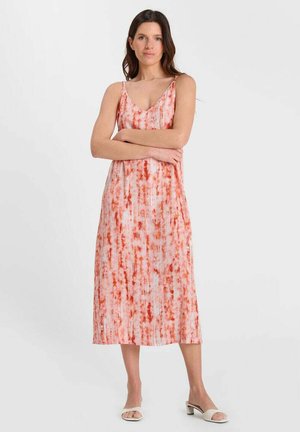 Vrouw staat met gekruiste armen en draagt een mouwloze midi-jurk met V-hals met een oranje-wit tie-dye patroon en witte open-toe blokhak sandalen.