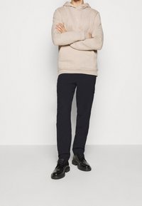 Sudadera beige con bolsillo canguro, pantalones cargo azul marino oscuros y zapatos negros de cordones. La sudadera presenta un pequeño logo blanco en el pecho.