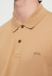 Άνδρας που φοράει μπεζ πόλο πουκάμισο με δύο μαύρα κουμπιά και το λογότυπο "BOSS" κεντημένο στο στήθος, εμφανιζόμενος από τον λαιμό έως το πάνω μέρος του κορμού.