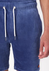 Shorts en tissu ribbed bleu avec une taille élastique et un cordon blanc. Dispose de poches latérales et d'un patch logo près de l'ourlet.
