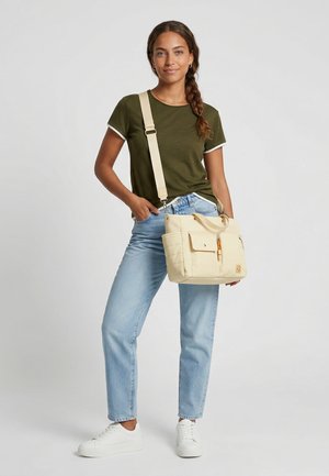 Vrouw met een olijfgroen t-shirt, lichtblauwe jeans en witte sneakers, met een crèmekleurige crossbodytas met voorvakken.