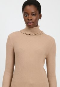 Donna con capelli corti che indossa un maglione beige a coste con colletto arricciato, piccoli orecchini a cerchio e trucco neutro.