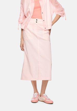 Woman wearing a light pink midi skirt, coral pink top, light pink jacket, and pink and white sneakers, standing with one hand in pocket.