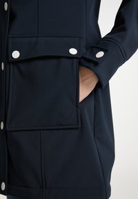 Granatowa kurtka softshell z zatrzaskowymi kieszeniami, mankietami na guziki i lekko dopasowanym krojem. Wykonana z gładkiego, teksturowanego materiału.