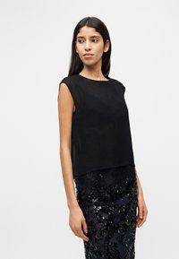 Jeune femme portant un haut noir transparent sans manches et une jupe noire ornée de sequins, debout devant un fond blanc uni.