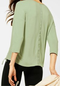Light green, three-quarter sleeve top with a scoop neckline, featuring a decorative stitch detail along the back and ties at the hem.