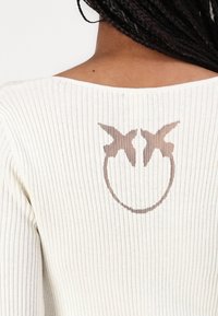 Off-white ribbattuupaja, jolla on leveä kaula-aukko, jossa on ruskea graafinen kuva kahdesta linnusta ja pyöreä muotoilu takana.