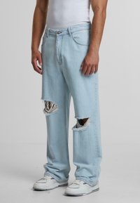 Light blue ripped jeans with large frayed knee openings and a relaxed fit. Paired with white sneakers.