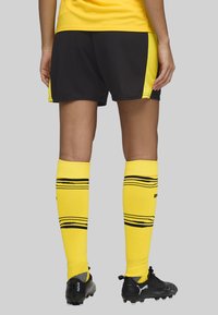 Calze da calcio gialle con strisce nere, abbinate a pantaloni corti neri con accenti gialli. Le calzature presentano scarpini da calcio neri.