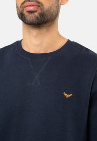 Donkerblauwe sweatshirt, van katoen, met ribgebreide ronde hals, voorzien van een oranje geborduurd logo op de linkerborst en kruissteekdetails op de halslijn.