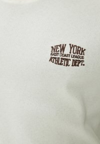 Lys grå ribbet sweater med brun broderet tekst: "NEW YORK EAST COAST LEAGUE ATHLETIC DEPT."