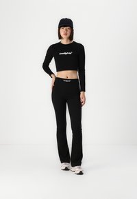 Black long-sleeve crop top with logo, paired with black flared pants. Features a fitted design. Model wears a black cap and light sneakers.