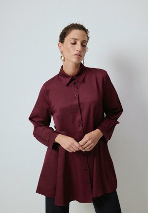 Γυναίκα με μαλλιά μαζεμένα πίσω, που φοράει χρυσά σκουλαρίκια κρεμαστά και μακρυμάνικο σκουρόχρωμο πουκάμισο βυσσινί με κουμπιά, στέκεται μπροστά σε ένα απλό φωτεινό φόντο.