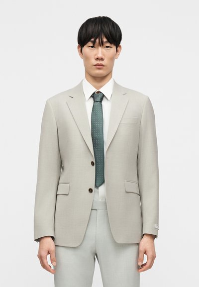 Γκρι σακάκι με εγκοπές στα πέτα και δύο μπροστινές τσέπες, συνδυασμένο με λευκό πουκάμισο και πράσινη γραβάτα με λευκές βούλες.