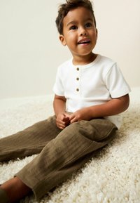 White short-sleeve henley shirt with three brown buttons; brown-textured pants. Child sitting on a soft, light-colored rug.