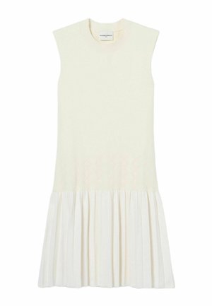 Joven mujer con vestido blanco plisado sin mangas, pañuelo rojo y azul al cuello, zapatillas oscuras y sosteniendo un pequeño bolso marrón.