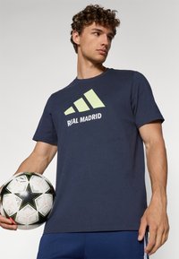 Marineblå bomulds t-shirt med korte ærmer, prydet med et lyst grønt Adidas-logo og teksten "REAL MADRID". Modellen holder en mønstret fodbold.