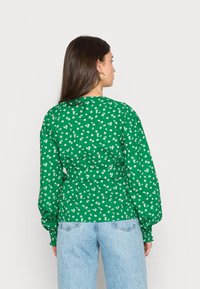 Blusa verde com padrão floral, com mangas bufantes e cintura ajustada. Tecido texturizado com pequenas flores brancas por toda a parte. Ajuste casual.