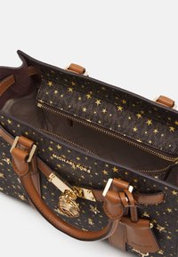 Mörkbrun Michael Kors-handväska med mönster av guldstjärnor, handtag av ljust läder, guldfärgad låsdekoration och öppet fack med dragkedja upptill.
