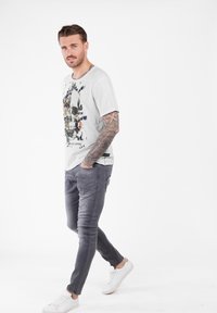 Mann mit ärmellangen Tätowierungen, der ein weißes T-Shirt mit floralem Totenkopf-Druck, graue Jeans und weiße Sneakers trägt und vor einem schlichten weißen Hintergrund geht.