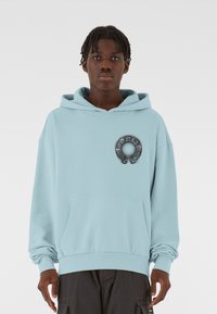 Lys blå hoodie lavet af blødt stof med en frontlomme, der har et rundt, metallisk logodetalje på brystet.