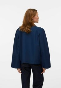 Marineblauwe cropped jas met brede mouwen, voorzien van schouderknopen en een gladde textuur. Gecombineerd met donkere spijkerbroeken.