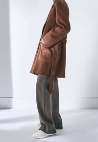 Cappotto in misto lana marrone con cintura e tasche laterali, abbinato a pantaloni grigi a gamba larga e sneakers bianche. Silhouette semplice e rilassata.