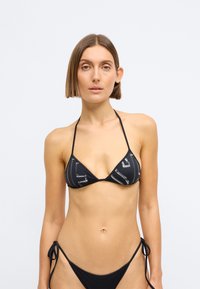 Μαύρο σετ μπικίνι που διαθέτει τοπ τριγωνικού σχήματος με γυαλιστερό σχέδιο και λεπτούς φιόγκους, συμπληρωμένο με αντίστοιχο κάτω μέρος με φιόγκους στο πλάι. Λείο υλικό.