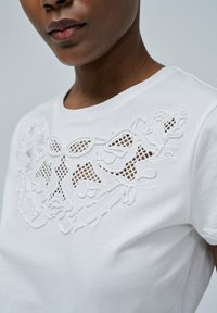 Camiseta blanca de algodón con intrincados bordados en blanco y patrones florales recortados en el escote. Diseño de manga corta.