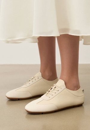 Pies con zapatos de cuero crema con cordones y suelas marrones, combinados con una falda blanca, de pie sobre un suelo beige.