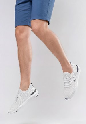 Person trägt weiße Stricksneaker mit schwarzen Akzenten und blaue Shorts, festgehalten im Sprung vor einem schlichten hellgrauen Hintergrund.