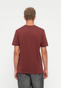 Mand med kort krøllet hår iført en ensfarvet maroon kortærmet t-shirt og mørkegrå jeans, stående mod en hvid baggrund og vendt væk.