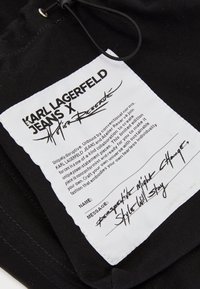 Musta kangastunniste, jossa on valkoista tekstiä. Sisältää brändin "KARL LAGERFELD JEANS X Atelier Reserve." Sisältää hoito-ohjeet ja viestiosion.
