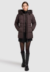 Giacca a vento marrone con cappuccio, chiusura con zip e cordini regolabili. Presenta un design trapuntato e due tasche laterali.