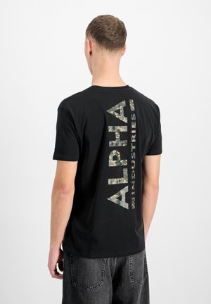 Person viewed from behind wearing a black t-shirt with large vertical camouflage "Alpha Industries" text and dark denim jeans.