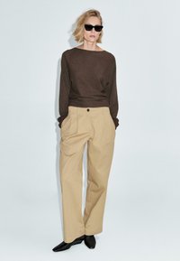 Donna che indossa occhiali da sole neri, maglione lungo marrone, pantaloni beige a vita alta e a gamba larga, e scarpe nere a punta su uno sfondo neutro.