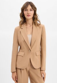 Femme avec des cheveux châtain clair ondulés, portant un blazer beige et un pantalon assorti, se tenant devant un fond blanc.