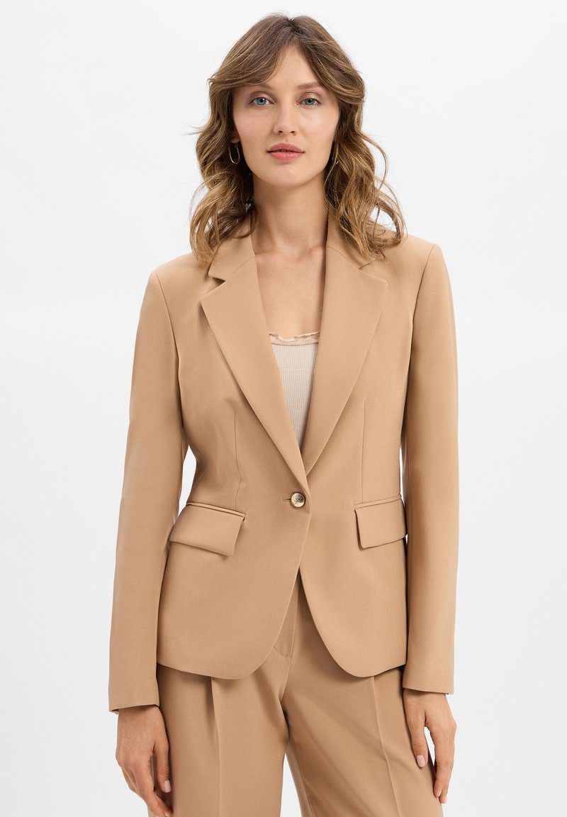 Femme avec des cheveux châtain clair ondulés, portant un blazer beige et un pantalon assorti, se tenant devant un fond blanc.