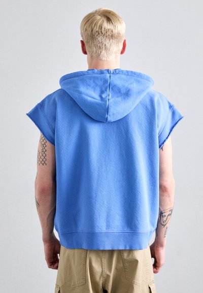 Blaues ärmelloses Kapuzensweatshirt mit lockerer Passform, strukturiertem Stoff, ausgefransten Kanten und geripptem Saum, kombiniert mit khakifarbenen Shorts.