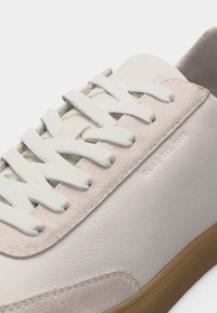 Κοντινό πλάνο λευκού δερμάτινου αθλητικού παπουτσιού με απαλές ροζ σουέντ λεπτομέρειες, λευκά κορδόνια και ελαφρώς καφέ λαστιχένια σόλα.