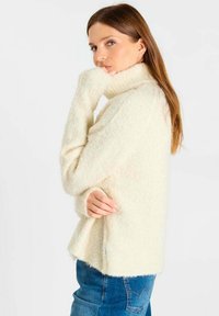 Vrouw met lang bruin haar, gekleed in een crèmekleurige, zachte coltrui en een blauwe spijkerbroek, kijkt over haar schouder.