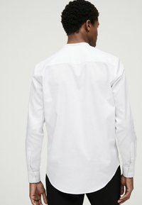 Hvid langærmet skjorte med lige snit, der har en rygsyede og knapmanchetter. Stoffet virker glat og let.