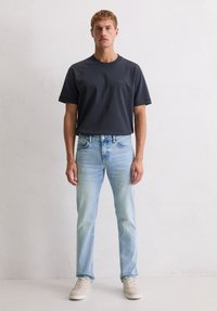 Jeune homme adossé à un mur blanc, portant un t-shirt gris foncé, un jean bleu clair et des baskets beiges, tourné vers l'avant.