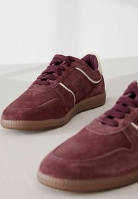 Wildleder Sneakers in Kastanienrot mit einer strukturierten Oberfläche, kontrastierenden cremefarbenen Akzenten an den Seiten und flachen Schnürsenkeln. Gummisohle und gepolsterter Schaft.