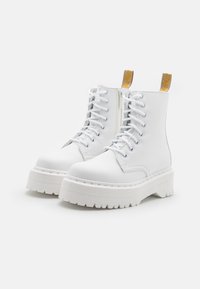 Botas de combate de plataforma blanca con suelas gruesas y estriadas, cordones, cremalleras laterales y tiradores amarillos con marca en la parte posterior.