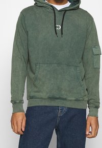 Sweat à capuche vert avec une finition délavée, poche avant et cordon de serrage réglable. Comprend une poche latérale et un logo discret sur la poitrine.