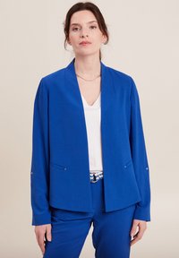 Blazer blu con maniche lunghe e collo sciallato, dotato di due tasche frontali e dettagli metallici, indossato sopra una maglietta bianca.