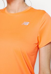 Camiseta deportiva naranja hecha de tela texturizada, con cuello redondo y un logo plateado de New Balance en el pecho izquierdo.