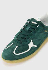 Baskets en daim vert avec des accents blancs, présentant un design perforé, des lacets et une semelle en caoutchouc naturel. Des coutures détaillées ajoutent de la texture.